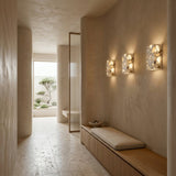 A serene, minimalist hallway with beige walls features a built-in wooden bench with cushions and the Terrano 1 Light Rectangular Travertine Wall Light in light beige casting soft natural light, leading to a room where a small tree is visible outside.