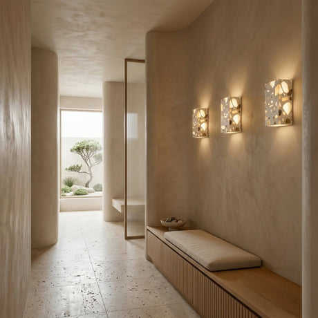 A serene, minimalist hallway with beige walls features a built-in wooden bench with cushions and the Terrano 1 Light Rectangular Travertine Wall Light in light beige casting soft natural light, leading to a room where a small tree is visible outside.