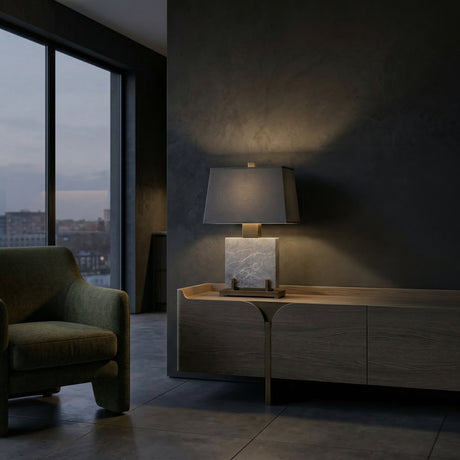 A modern, dimly lit living room with a green armchair, wooden sideboard, and the Karsen 1 Light Framed Marble Table Lamp in Brass & Black casting warm light against a dark gray wall by large windows overlooking the city at dusk.