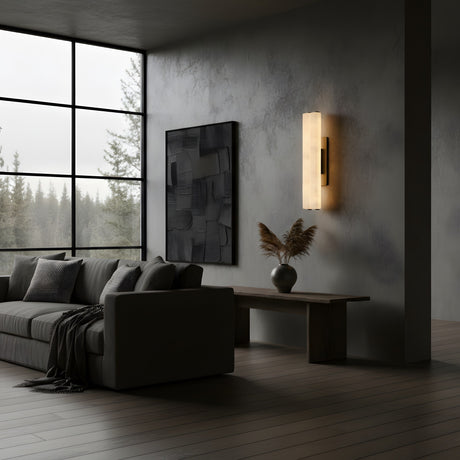 A modern living room features a gray sofa, neutral pillows, textured wall, minimalist painting, wooden bench with dried grass vase, the Selvara LED Large Alabaster Double Wall Light in Matte Black & Soft White softly glowing, and large windows with a view of trees.