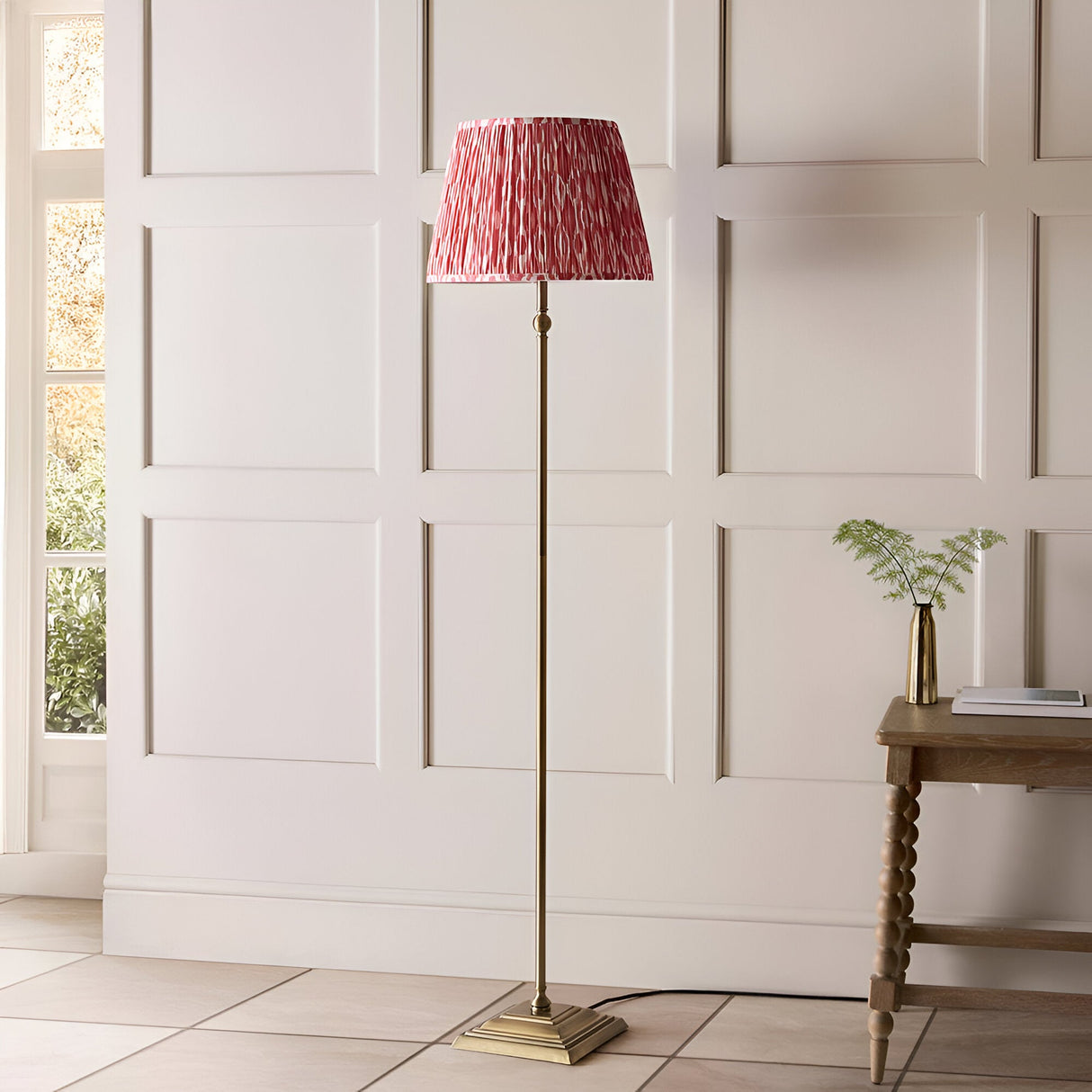 Endon Caldmere floor lamp with antique brass stem and stepped base, topped with a pleated coral pink Ikat fabric shade, in a panelled hallway.