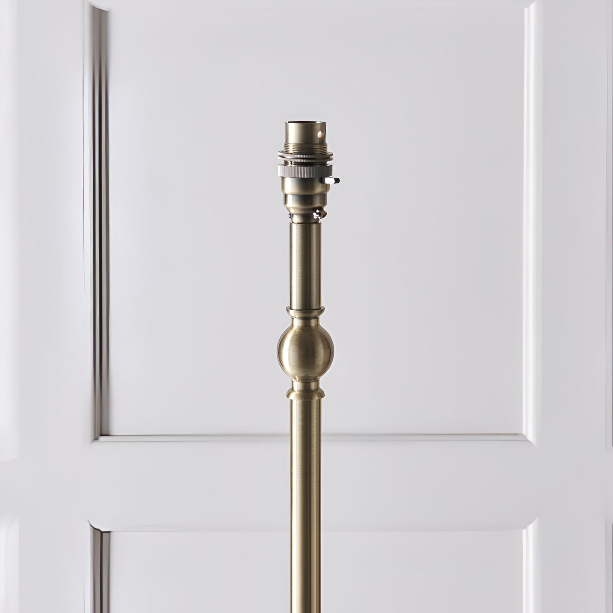Close-up of the Endon Caldmere floor lamp's antique brass stem with B22 lamp holder and turned ball detail, against a white panelled door.