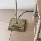 Close-up of the Endon Caldmere floor lamp's stepped square antique brass base and slim stem beside a wooden bobbin-legged stool.
