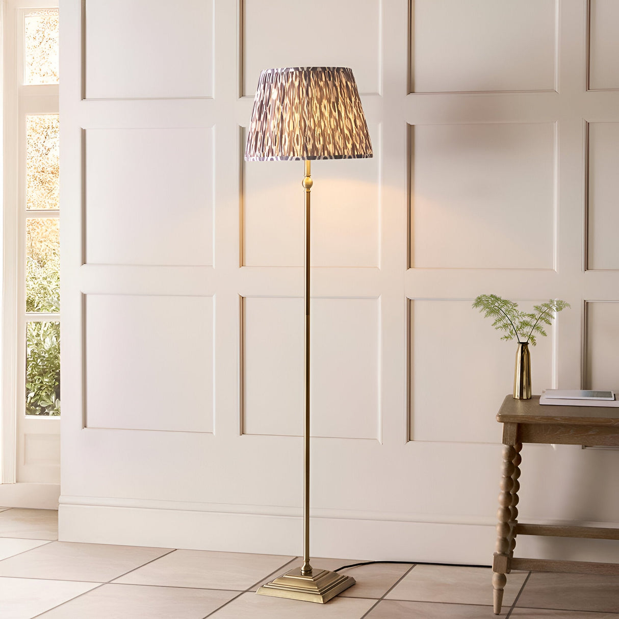 Caldmere Ikat Floor Lamp in Grey, with antique brass metal stem and square base, pleated grey patterned fabric shade, shown in a panelled living room setting.