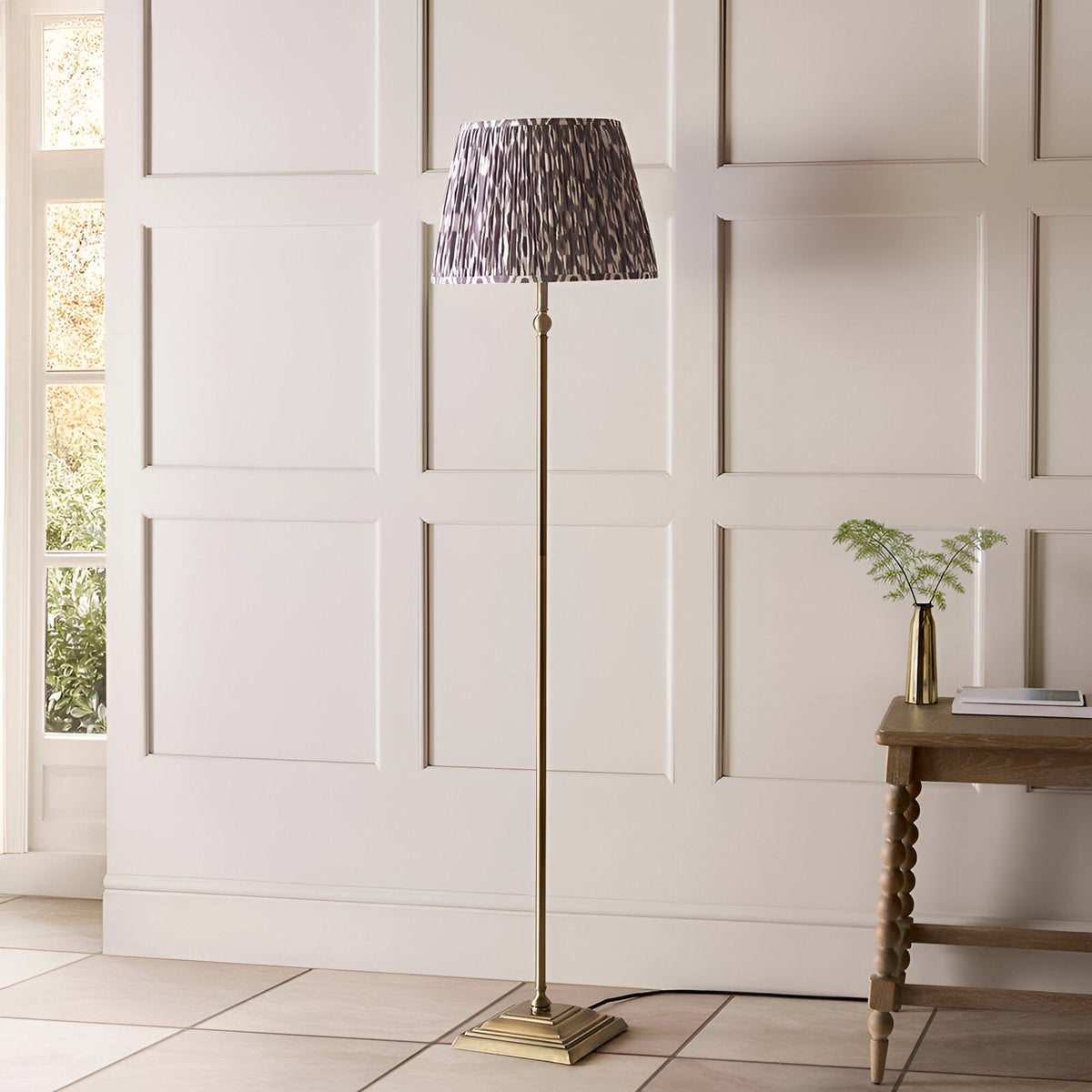 Caldmere Ikat Floor Lamp in Grey with an antique brass metal stand, square base, and a pleated grey patterned fabric shade, shown in a panelled living room.
