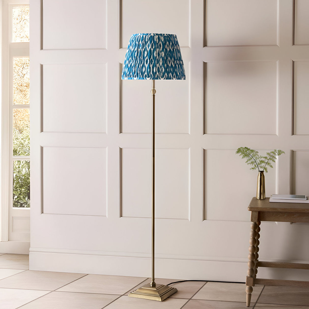Endon Caldmere floor lamp with antique brass stem and stepped base, topped by a pleated marlin blue Ikat shade, in a panelled hallway setting.