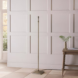 Endon Caldmere floor lamp base in antique brass with stepped square footing and B22 holder, shown without shade beside a wooden console in a panelled room.