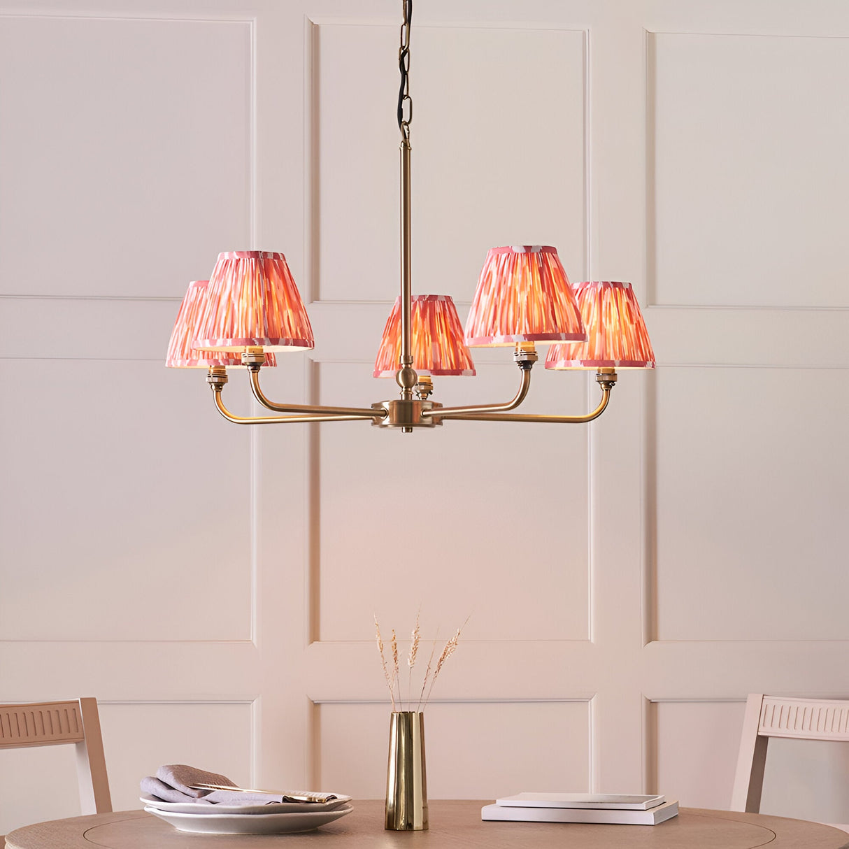 Endon Caldmere 5 light pendant with antique brass arms and coral pink pleated ikat tapered shades, hung above a dining table against a panelled wall.