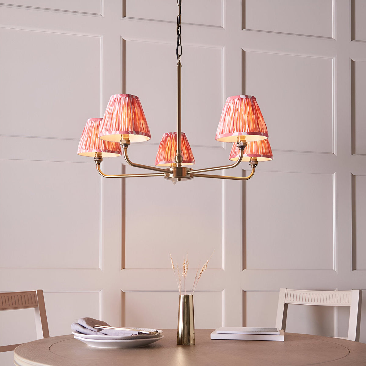 Endon Caldmere 5 light pendant with antique brass arms and coral pink pleated ikat tapered shades, hung above a dining table against panelled walls.