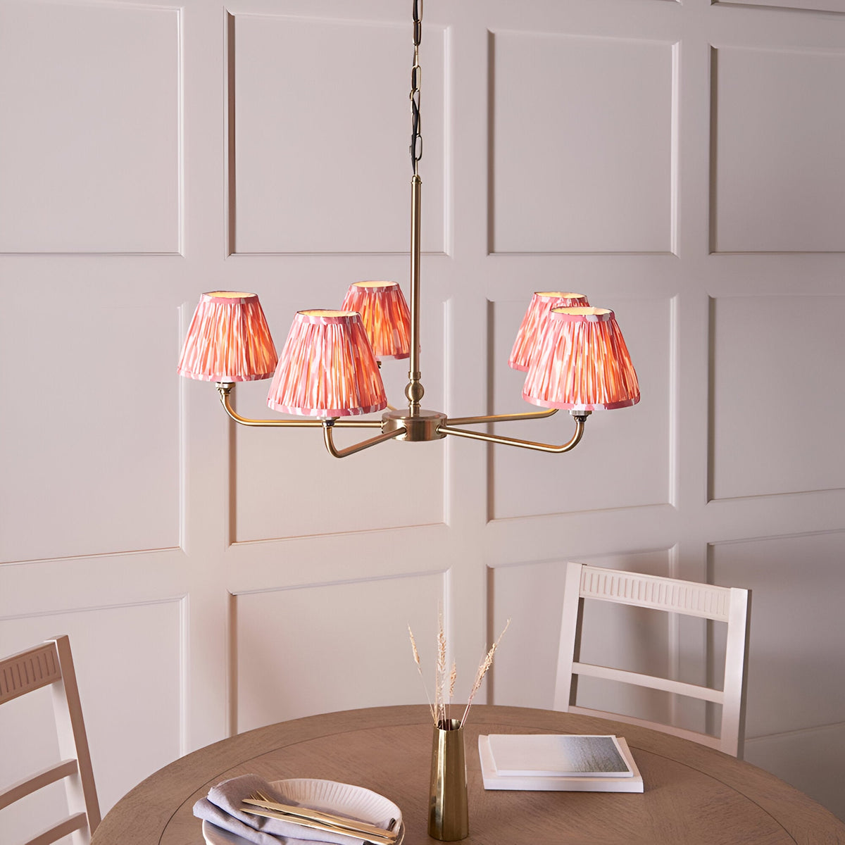 Endon Caldmere five light pendant with antique brass arms and coral pink pleated ikat tapered shades, hung above a wooden dining table.