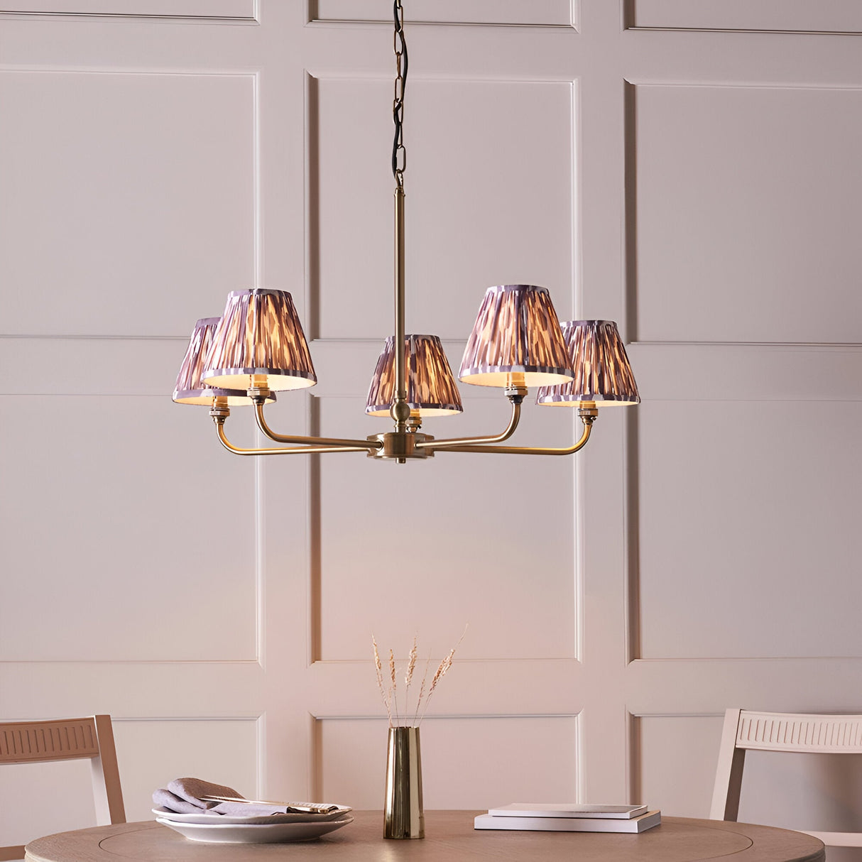 Caldmere 5 Light Ikat Pendant in antique brass with grey pleated fabric shades, shown hanging above a dining table in a panelled room.