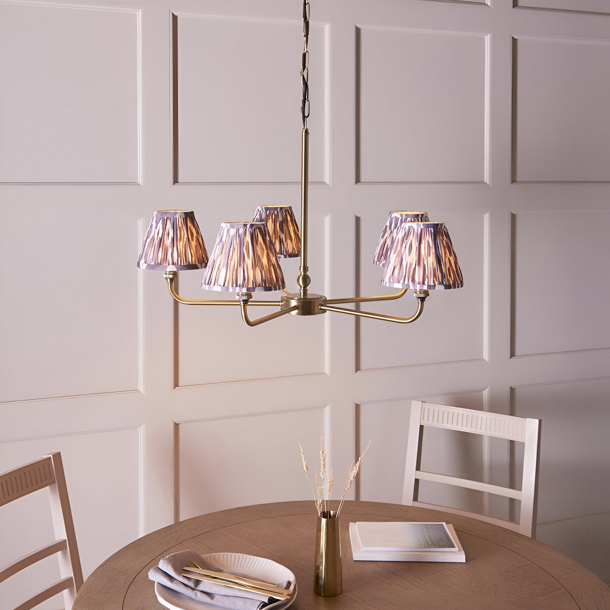 Caldmere 5 Light Ikat Pendant in antique brass with grey pleated fabric shades, hung above a dining table in a panelled room.