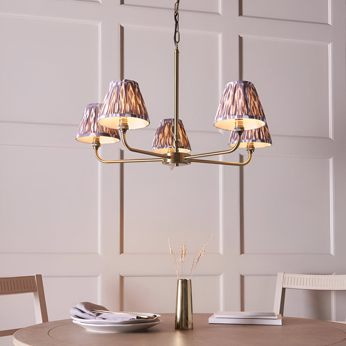 Caldmere 5 Light Ikat Pendant in antique brass with grey pleated fabric tapered shades, hanging above a dining table in a light-panelled room.