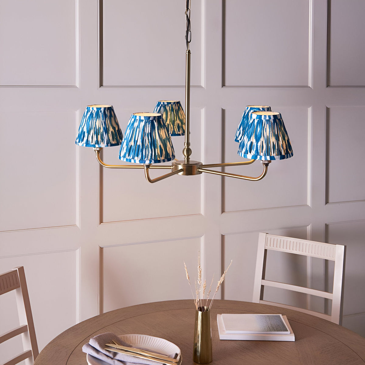 Endon Caldmere 5 light pendant with tapered pleated marlin blue ikat fabric shades on antique brass arms, shown above a dining table.