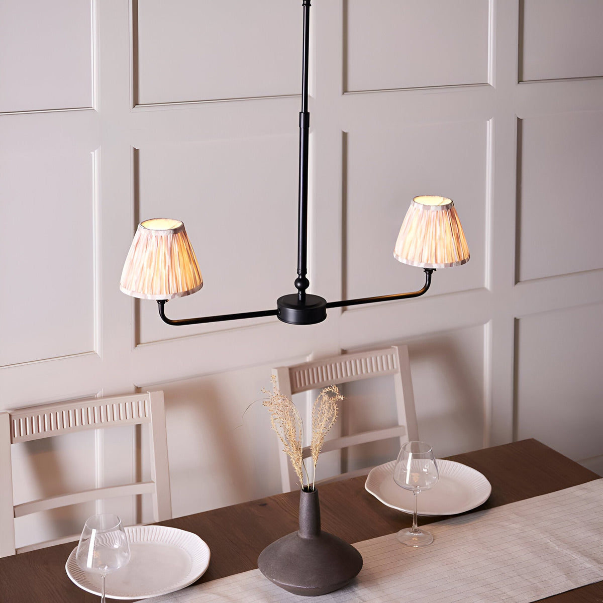 Endon Caldmere 2 light pendant with matt black linear frame and natural pleated tapered shades, shown above a dining table against panelled walls.