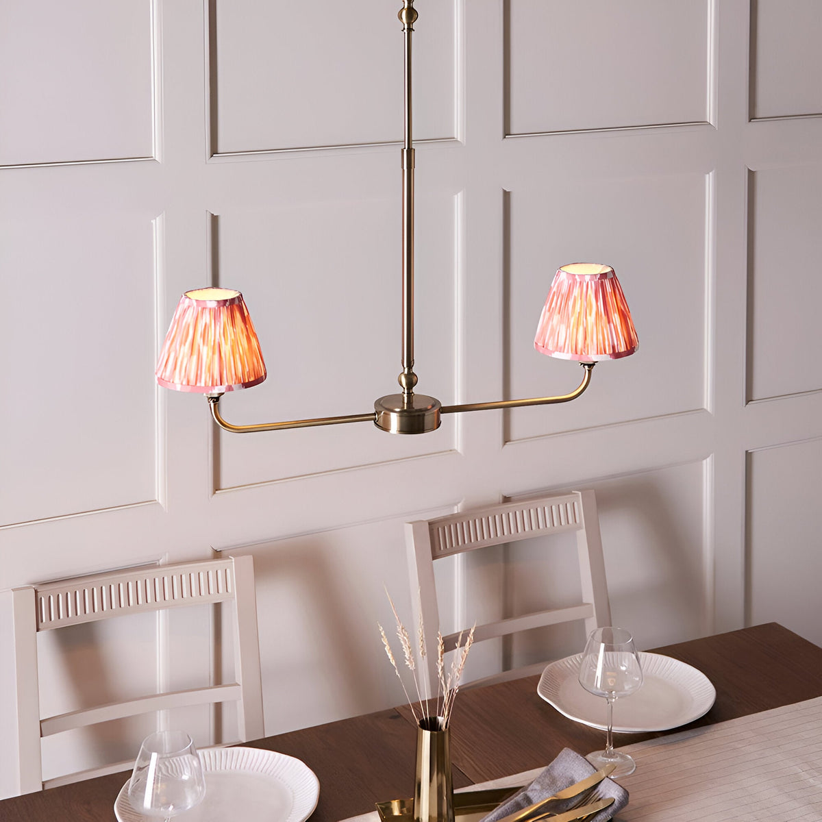 Endon Caldmere 2 light linear pendant in antique brass with coral pink pleated ikat tapered shades, shown above a wood dining table.