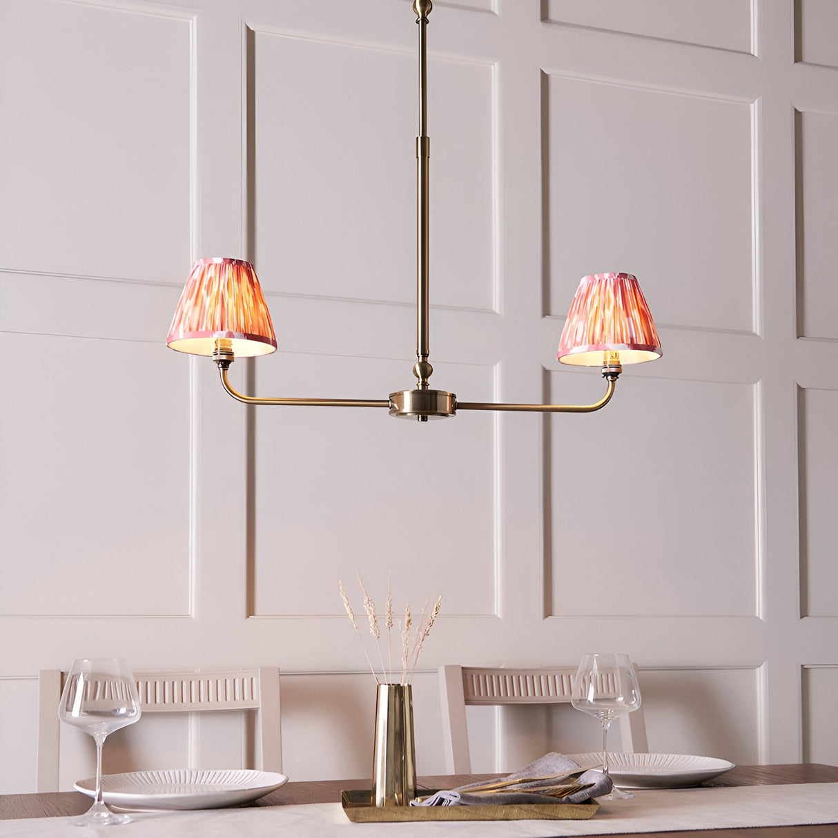 Endon Caldmere 2 light linear pendant in antique brass with coral pink pleated ikat shades, hung above a dining table against panelled walls.