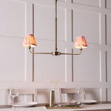 Endon Caldmere 2 light linear pendant in antique brass with coral pink pleated ikat shades, hung above a dining table against panelled walls.