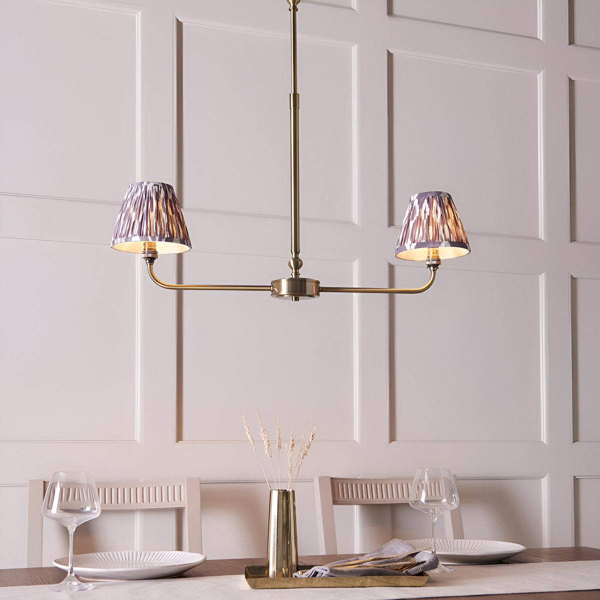 Caldmere 2 Light Ikat Pendant in antique brass with grey pleated tapered shades, shown above a dining table in a panelled room.