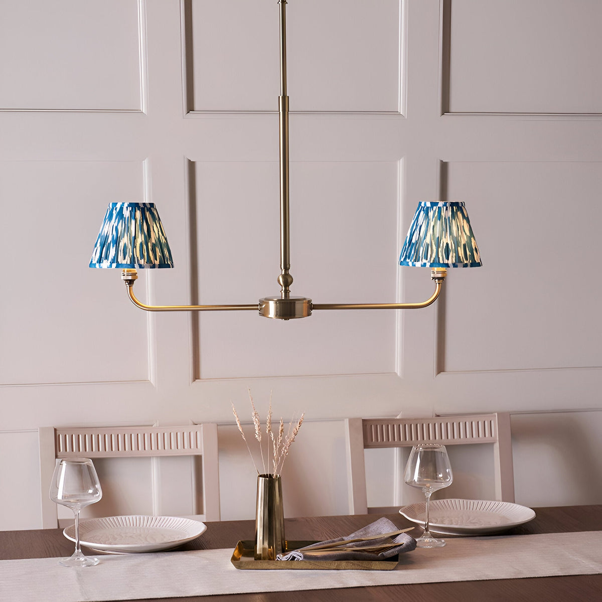 Endon Caldmere 2 light pendant in antique brass with twin tapered blue ikat pleated shades on a linear frame, shown above a dining table.