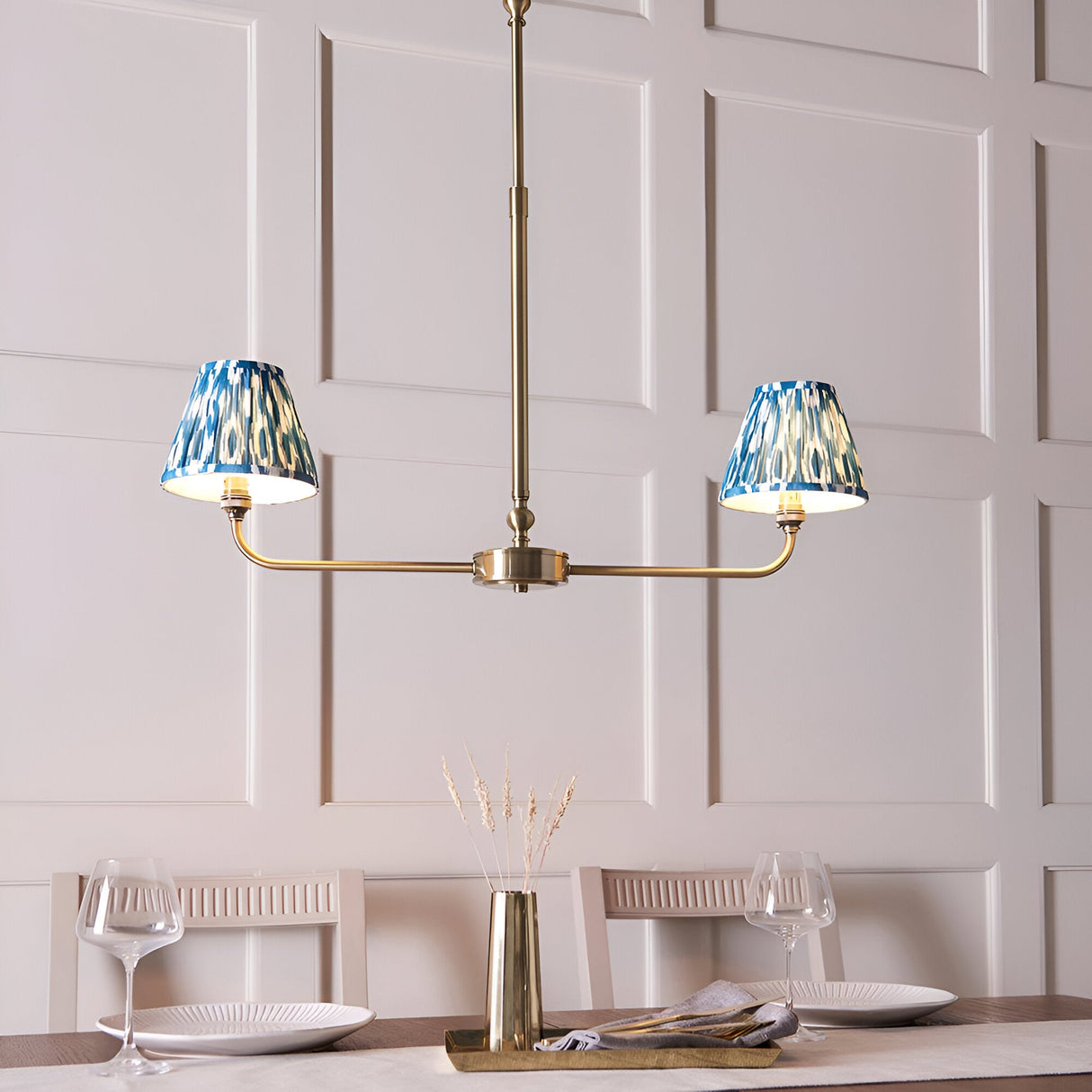 Endon Caldmere 2 light pendant with tapered blue ikat fabric shades on an antique brass linear frame, shown above a dining table.
