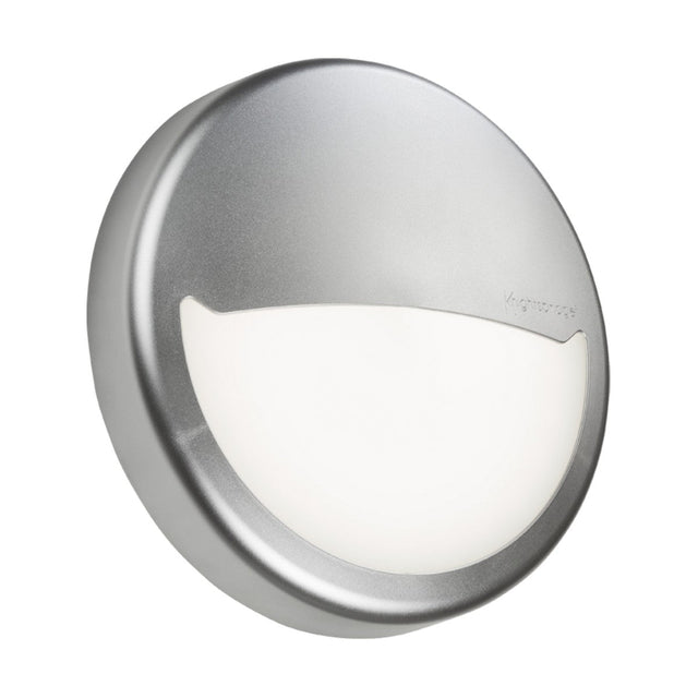 A round, metallic BT14 wall light fixture with a frosted white cover is elegantly complemented by the Eyelid Accessory For BT14 in Silver. Designed for sleek and modern aesthetics, it features a partial metal casing and an exposed semicircle that emits light.
