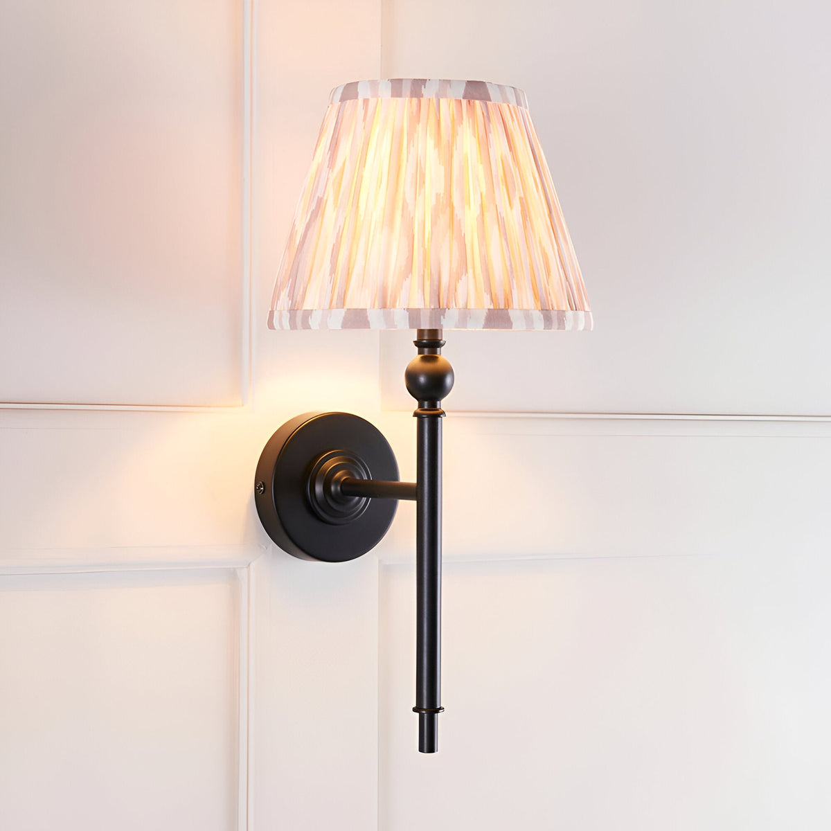 Endon Caldmere wall light with matt black metalwork and a tapered pleated Ikat shade in beige tones, illuminated against a white panelled wall.