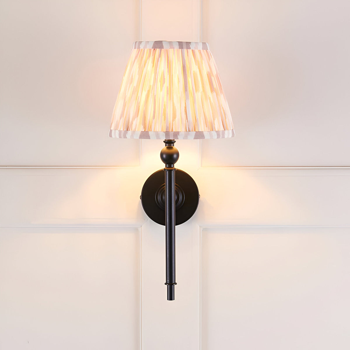 Endon Caldmere wall light with a tapered pleated beige Ikat fabric shade on slim matt black metalwork, mounted on a white panelled wall.