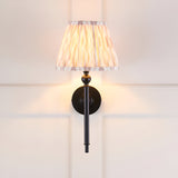 Endon Caldmere wall light with a tapered pleated beige Ikat fabric shade on slim matt black metalwork, mounted on a white panelled wall.