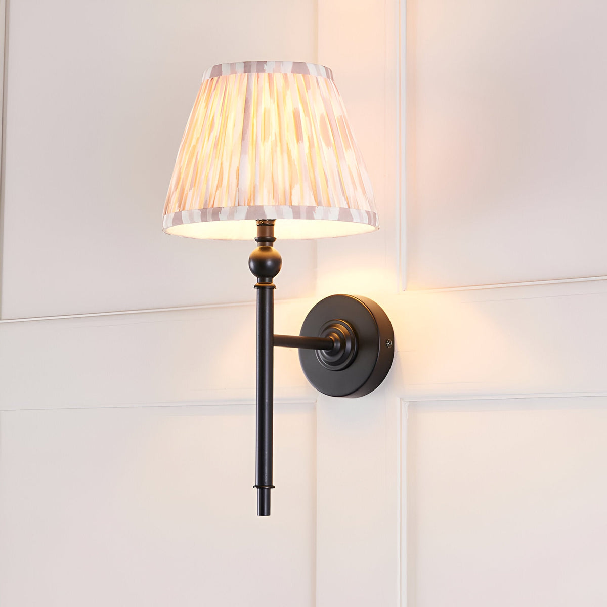 Endon Caldmere wall light with matt black metalwork and a tapered hand-pleated beige Ikat fabric shade, mounted on a white panelled wall.