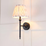 Endon Caldmere wall light with matt black metalwork and a tapered hand-pleated beige Ikat fabric shade, mounted on a white panelled wall.