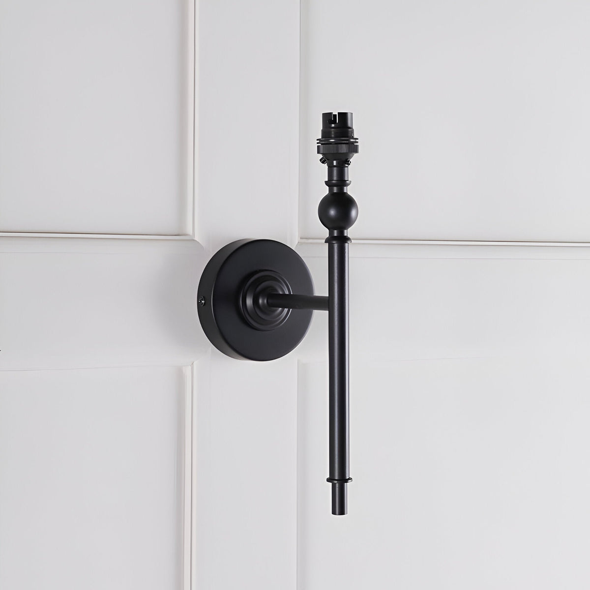 Endon Caldmere wall light base in matt black with slim upright stem, ball detail and B22 lamp holder, mounted on white panelled wall, shade not fitted.