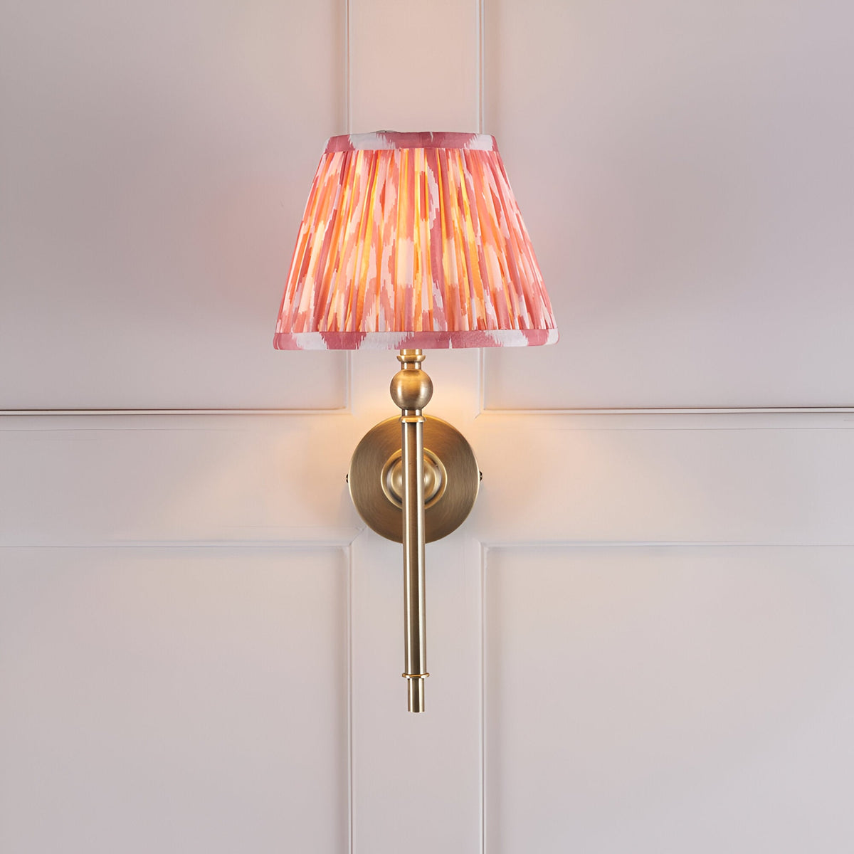 Caldmere Ikat Wall Light in Coral, featuring a pleated coral pink tapered fabric shade on an antique brass wall fitting, mounted on a panelled white wall.
