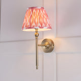 Caldmere Ikat Wall Light in Coral, featuring a pleated coral pink tapered fabric shade on an antique brass wall bracket, mounted on a light panelled wall.
