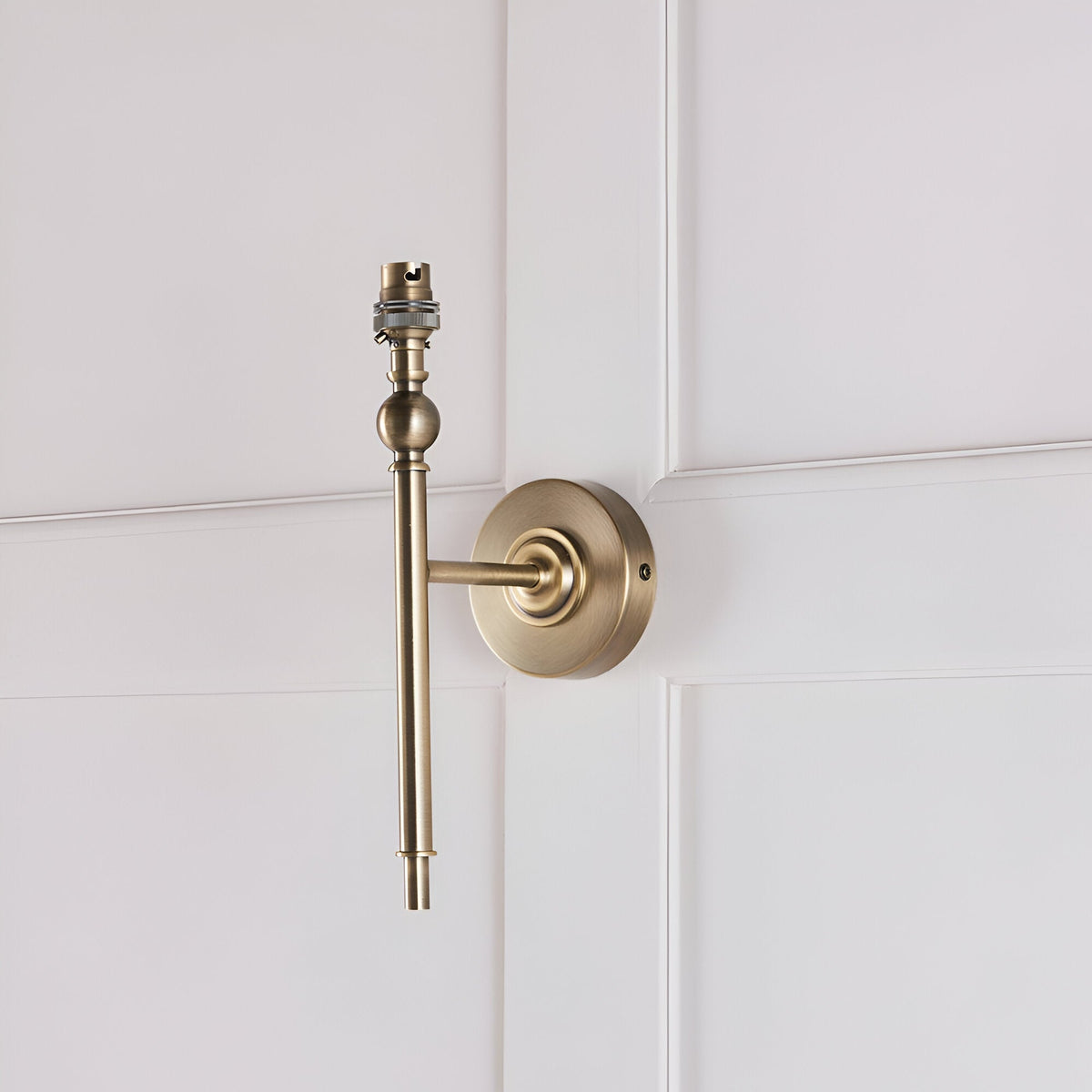 Caldmere Ikat Wall Light in antique brass, shown without its shade, mounted on a light panelled wall with a round backplate and slim upright arm.