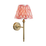 Caldmere Ikat Wall Light in coral pink with a pleated tapered fabric shade on an antique brass arm and round wall mount, shown on a plain white background.