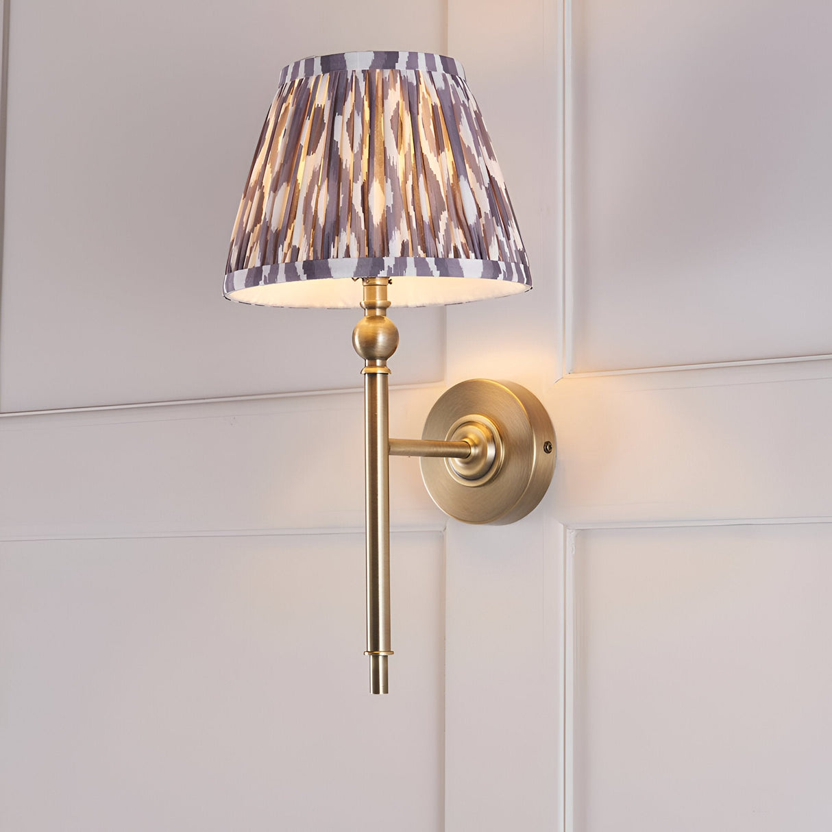 Endon Caldmere Ikat wall light in antique brass with a tapered pearl grey pleated Ikat shade, mounted on a white panelled wall.