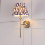 Endon Caldmere Ikat wall light in antique brass with a tapered pearl grey pleated Ikat shade, mounted on a white panelled wall.