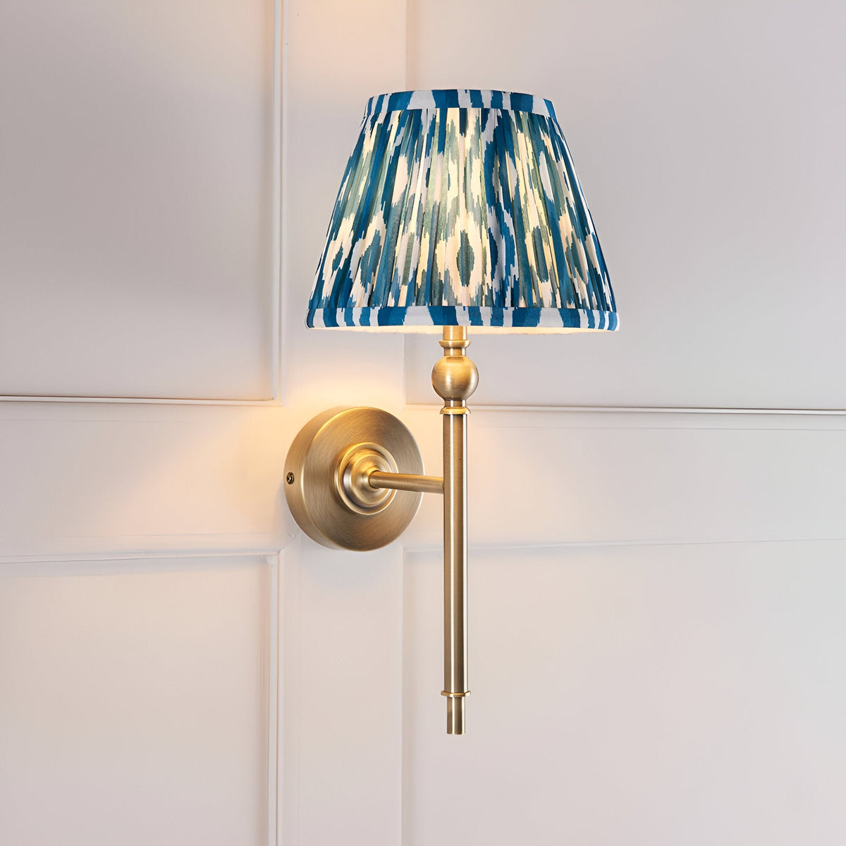 Caldmere Ikat Wall Light in antique brass with a pleated blue patterned fabric tapered shade, mounted on a wall-panelled background.