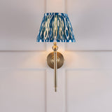 Caldmere Ikat Wall Light in Blue with a pleated tapered fabric shade on an antique brass mount, shown against a light panelled wall.