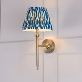 Caldmere Ikat Wall Light in Blue with pleated tapered fabric shade on an antique brass arm and round wall mount, shown against a panelled white wall.