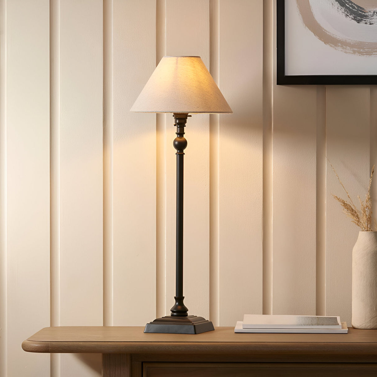 Endon Caldmont table lamp in matt black with a tall candlestick column and tapered natural linen shade on a wooden console beside framed art.
