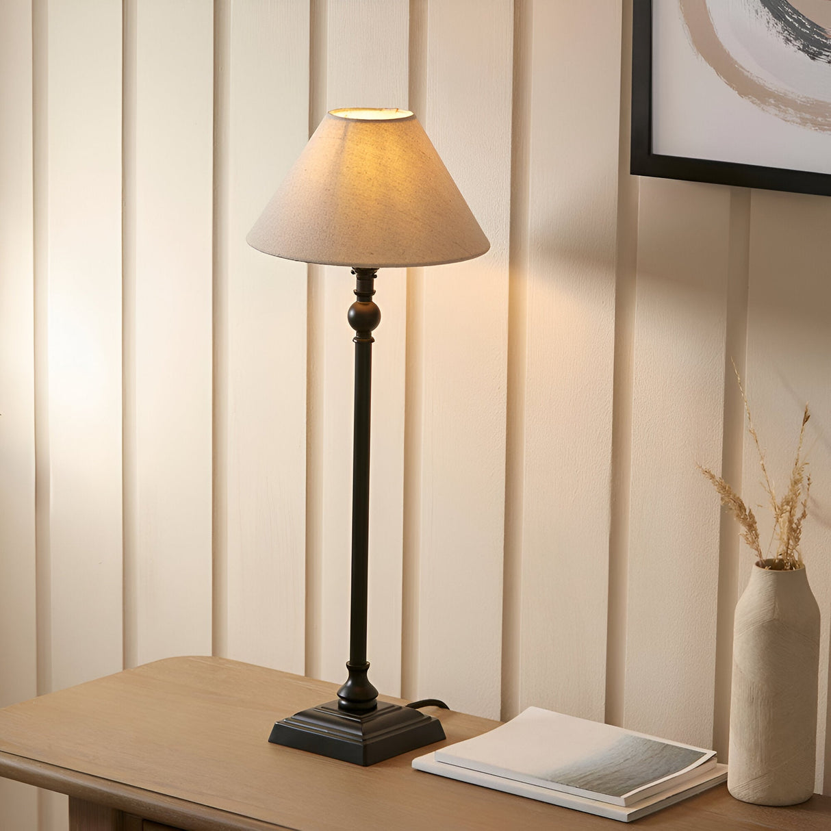 Endon Caldmont table lamp with slim matt black candlestick column and tapered natural linen shade, lit on a wood console beside a vase and books.