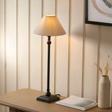 Endon Caldmont table lamp with slim matt black candlestick column and tapered natural linen shade, lit on a wood console beside a vase and books.