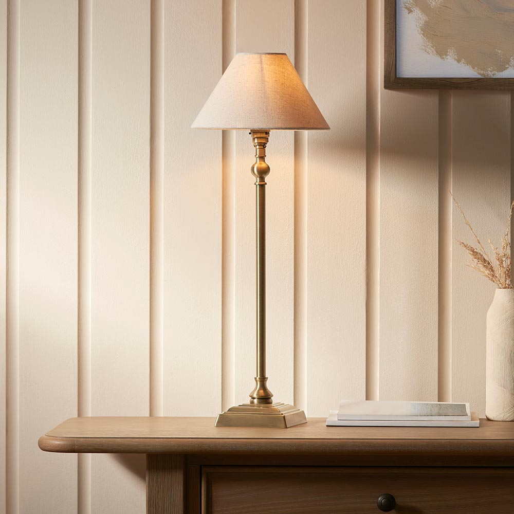Endon Caldmont candlestick table lamp in antique brass with tapered natural linen shade, lit on a wood console beside a vase in a panelled room.