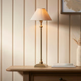 Endon Caldmont candlestick table lamp in antique brass with tapered natural linen shade, lit on a wood console beside a vase in a panelled room.