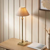 Endon Caldmont table lamp in antique brass with a slim candlestick column and tapered natural linen shade, lit on a wooden side table by a panelled wall.