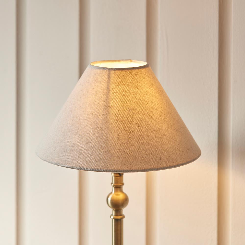 Endon Caldmont table lamp with antique brass candlestick base and tapered natural linen shade, lit against a cream panelled wall.