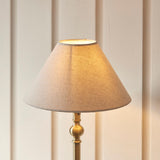 Endon Caldmont table lamp with antique brass candlestick base and tapered natural linen shade, lit against a cream panelled wall.