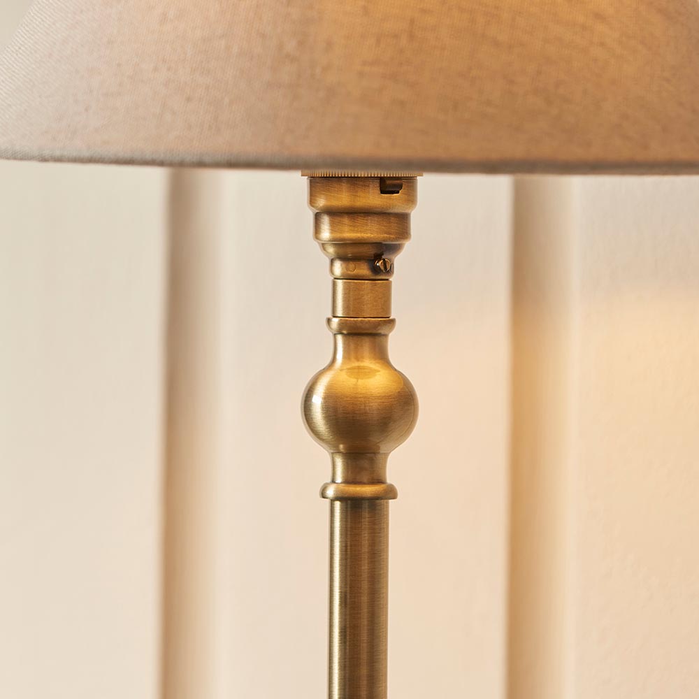 Close-up of the Endon Caldmont antique brass table lamp showing the turned candlestick column, lamp holder detail, and base of the natural linen shade.